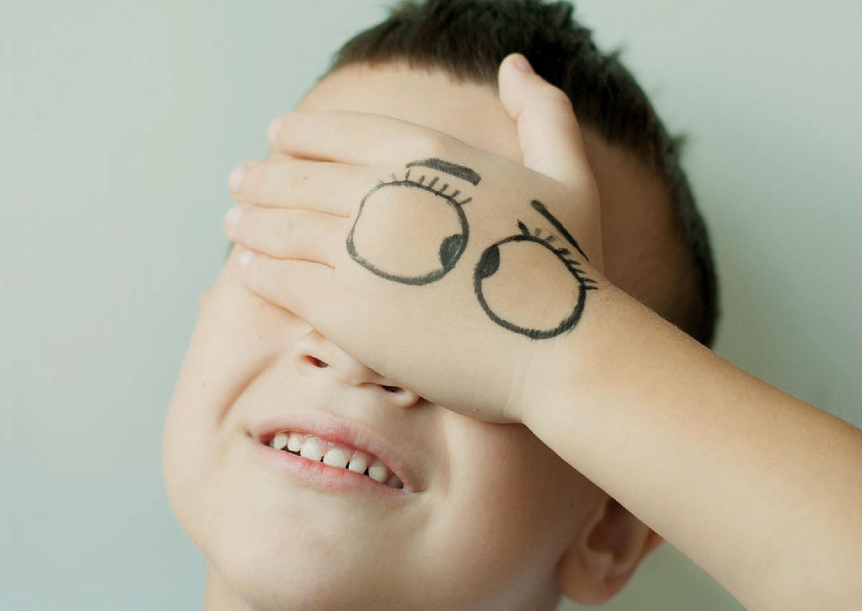 ein Junge hat seine Hand vor den Augen, auf der zwei schielende große Augen mit Augenbrauen aufgezeichnet sind