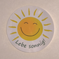 runder weißer Aufkleber mit lachender Sonne und der Aufschrift in Grün 'Lebe sonnig!'