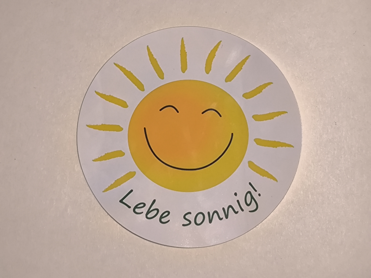 runder weißer Aufkleber mit lachender Sonne und der Aufschrift in Grün 'Lebe sonnig!'